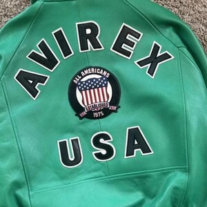 Puede incluir: Chaqueta de cuero verde con la palabra "AVIREX" en letras blancas en la parte superior de la espalda. Debajo, un parche circular con las palabras "ALL AMERICANS ESTABLISHED 1975" y un dise&ntilde;o de bandera estadounidense. La palabra "USA" est&aacute; debajo.