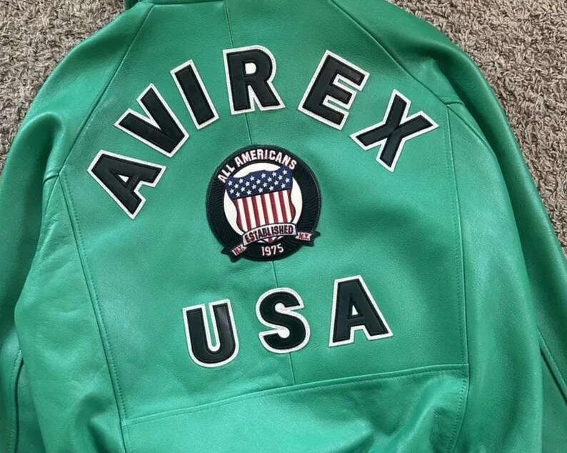Puede incluir: Chaqueta de cuero verde con la palabra "AVIREX" en letras blancas en la parte superior de la espalda. Debajo, un parche circular con las palabras "ALL AMERICANS ESTABLISHED 1975" y un dise&ntilde;o de bandera estadounidense. La palabra "USA" est&aacute; debajo.