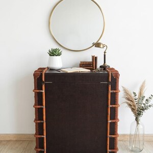 May include: A dark brown trunk-style cabinet with light brown trim and metal accents. A round gold mirror hangs above the cabinet, which is decorated with a succulent in a white pot, books, a lamp, and keys. A clear vase with dried plants is on the right.