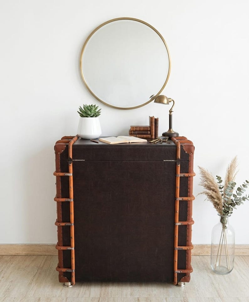 May include: A dark brown trunk-style cabinet with light brown trim and metal accents. A round gold mirror hangs above the cabinet, which is decorated with a succulent in a white pot, books, a lamp, and keys. A clear vase with dried plants is on the right.