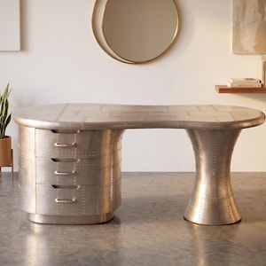 May include: A silver-toned metal desk featuring a curved, aeroplane-inspired design with visible rivet detailing. The desk includes a pedestal with three stacked drawers on the left side and a single flared, cylindrical support leg on the right side.