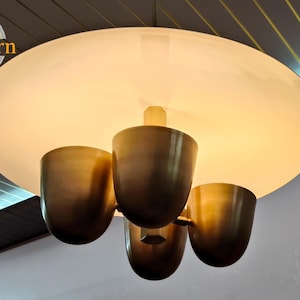 Op de afbeelding: Een moderne plafondlamp met een grote, ronde, matwitte kap. Vier bronskleurige, bekervormige lampen hangen eronder. De armatuur is gemonteerd op een donker houten plafond. Het SuziFurn-logo staat linksboven.