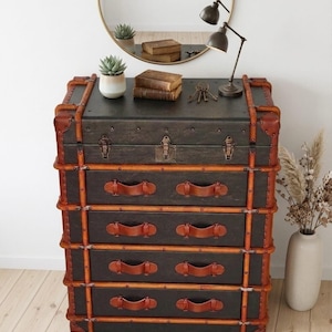May include: A vintage-style chest of drawers with a dark gray finish, accented with brown leather trim and handles. The chest has four drawers and is trimmed with wooden slats. A small lamp, mirror, and decorative items are on top.