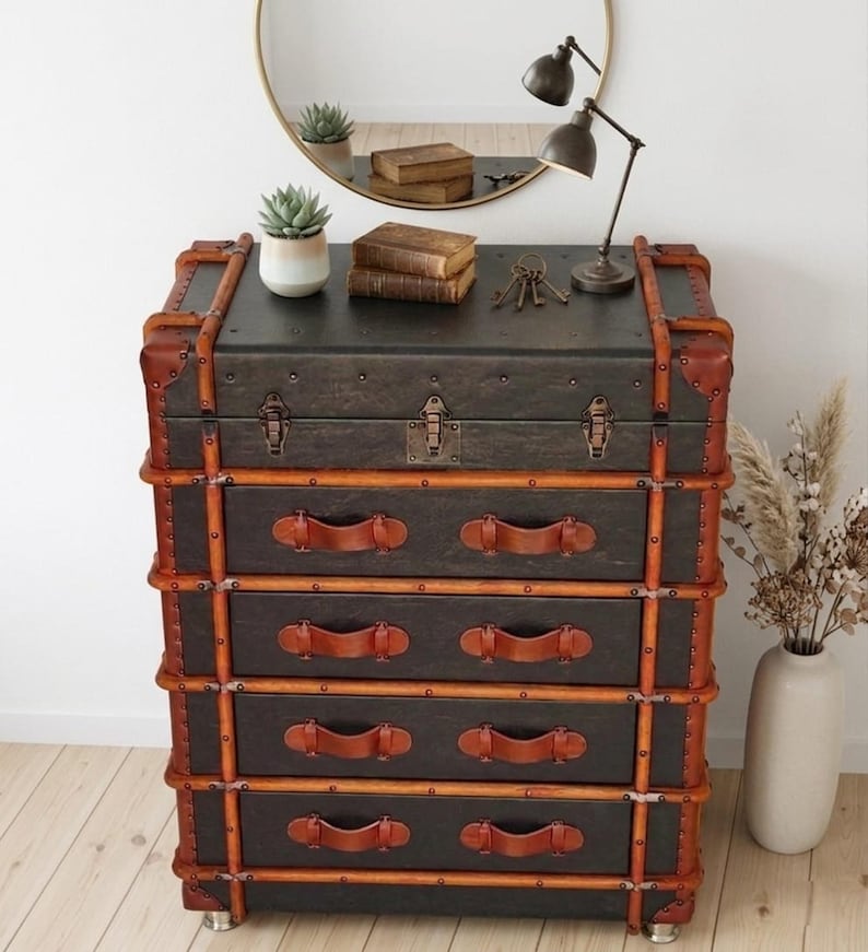 May include: A vintage-style chest of drawers with a dark gray finish, accented with brown leather trim and handles. The chest has four drawers and is trimmed with wooden slats. A small lamp, mirror, and decorative items are on top.