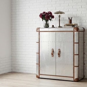 May include: A silver cabinet with brown leather trim and handles. The cabinet has two doors and is decorated with rivets. A lamp, books, and a vase of flowers sit on top. The cabinet is in a room with a white brick wall.