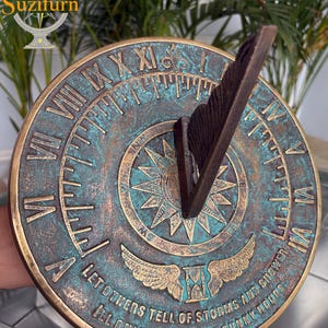 May include: A round, antique-style sundial with a weathered teal and bronze finish. Roman numerals mark the hours, and the center features a starburst design. The sundial has the text "LET OTHERS TELL OF STORMS AND SHOWER, I'LL ONLY MARK YOUR SUNNY HOURS."