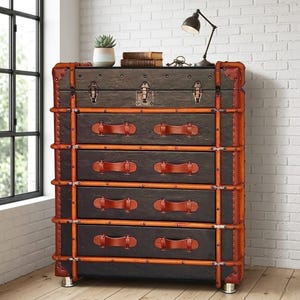May include: A dark brown chest of drawers with a vintage trunk design. It features leather handles and trim, metal accents, and a small lamp on top. The chest has five drawers and is set against a white brick wall.