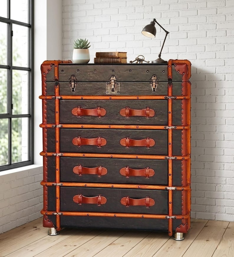 May include: A dark brown chest of drawers with a vintage trunk design. It features leather handles and trim, metal accents, and a small lamp on top. The chest has five drawers and is set against a white brick wall.
