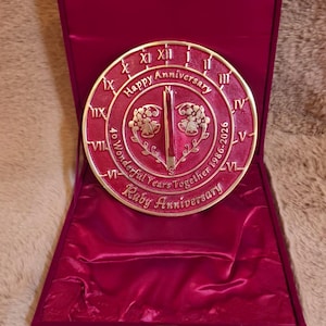 Könnte beinhalten: Eine kreisförmige, rubinrote Gedenkplakette mit einem zentralen Gnomon in einer mit Samt ausgekleideten Schatulle. Die Plakette zeigt römische Ziffern, den Text Happy Anniversary, 40 Wonderful Years Together 1986-2026, Ruby Anniversary und Glocken.