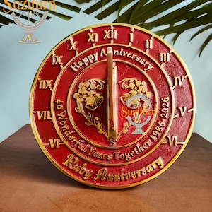 Puede incluir: Un reloj de sol rojo y dorado de aniversario de rubí con números romanos y el texto "Happy Anniversary" y "40 Wonderful Years Together 1986-2026". El reloj de sol tiene un gnomon central y elementos florales decorativos.