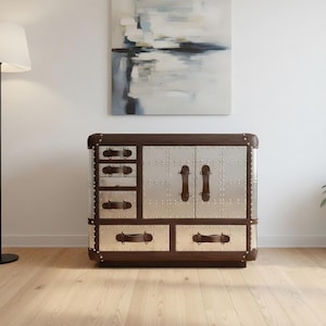 Puede incluir: Un mueble marrón y plateado con paneles metálicos remachados y asas de cuero. El mueble tiene cajones y puertas, y está enmarcado en madera oscura. También se ven una lámpara de pie y una planta en maceta.