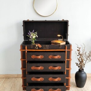 May include: A dark brown trunk-style dresser with four drawers, accented with tan leather handles and trim. The top is open, revealing a dark interior. A gold-framed mirror hangs above, with a vase of flowers and a black vase to the side.
