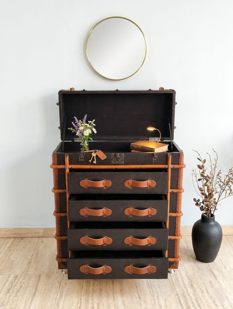 May include: A dark brown trunk-style dresser with four drawers, accented with tan leather handles and trim. The top is open, revealing a dark interior. A gold-framed mirror hangs above, with a vase of flowers and a black vase to the side.