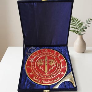 Pode incluir: Uma placa vermelha e dourada de aniversário de rubi numa caixa de veludo azul. A placa apresenta o texto "Happy Anniversary" e "Ruby Anniversary" com os anos 1986-2026. A caixa está aberta, revelando a placa e uma peça decorativa dourada.