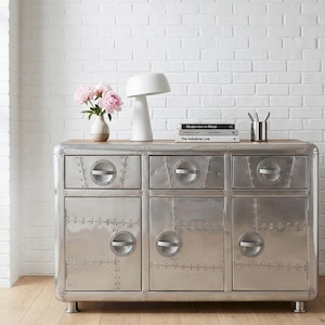 Puede incluir: Un mueble metálico plateado con seis cajones y puertas, con detalles remachados y tiradores circulares. El mueble tiene una tapa de madera y se apoya sobre patas cortas plateadas. Una lámpara blanca, libros y un jarrón con flores rosas encima.