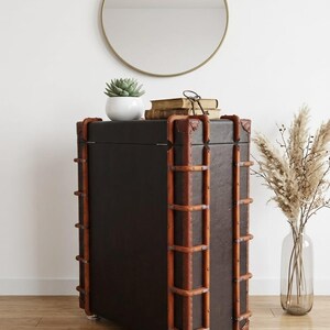May include: A dark brown trunk-style cabinet with brown wooden accents and metal rivets. A round gold mirror hangs on the white wall above the cabinet. A small white potted succulent and books sit on top of the cabinet.