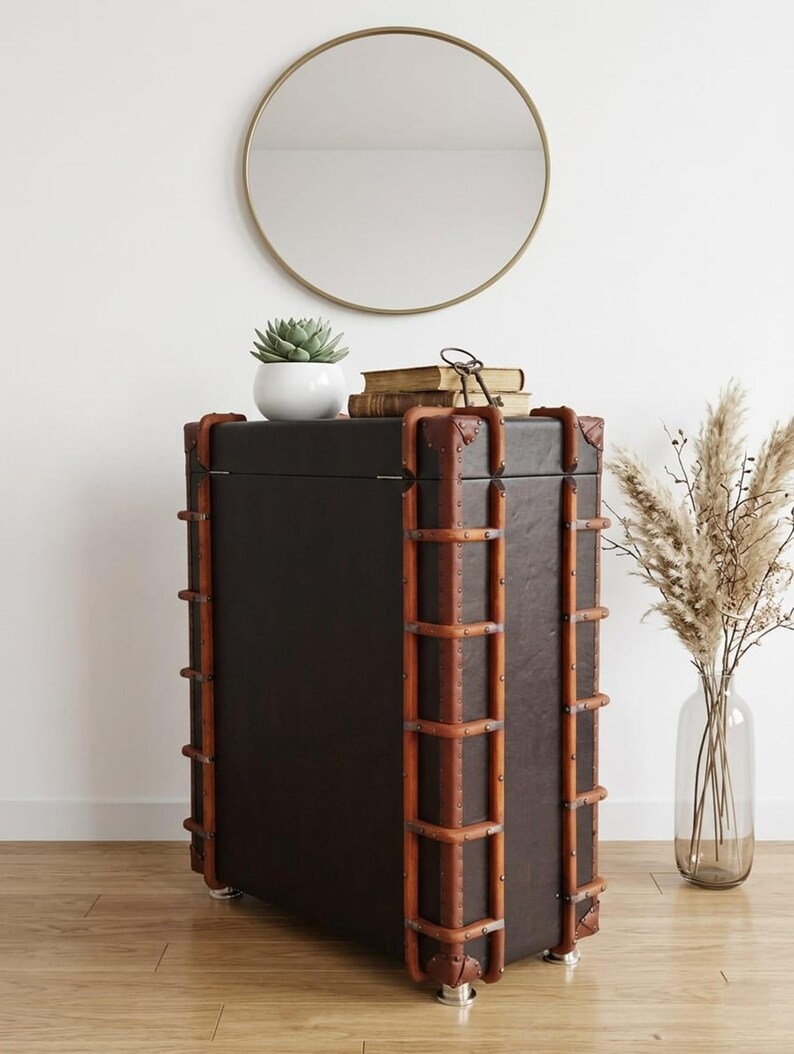 May include: A dark brown trunk-style cabinet with brown wooden accents and metal rivets. A round gold mirror hangs on the white wall above the cabinet. A small white potted succulent and books sit on top of the cabinet.