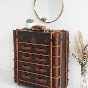May include: A dark brown chest of drawers with five drawers, accented with tan leather straps and handles. The chest has a vintage trunk design, with metal corner protectors and a lock. A round gold mirror hangs above.