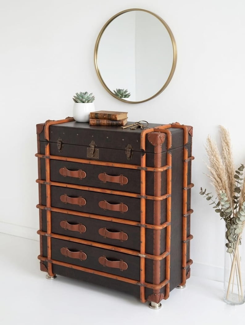 May include: A dark brown chest of drawers with five drawers, accented with tan leather straps and handles. The chest has a vintage trunk design, with metal corner protectors and a lock. A round gold mirror hangs above.