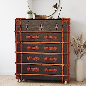 May include: A dark gray chest of drawers with five drawers, trimmed with brown leather and metal accents. The chest has brown leather handles and metal corner protectors. The chest is on silver metal feet.
