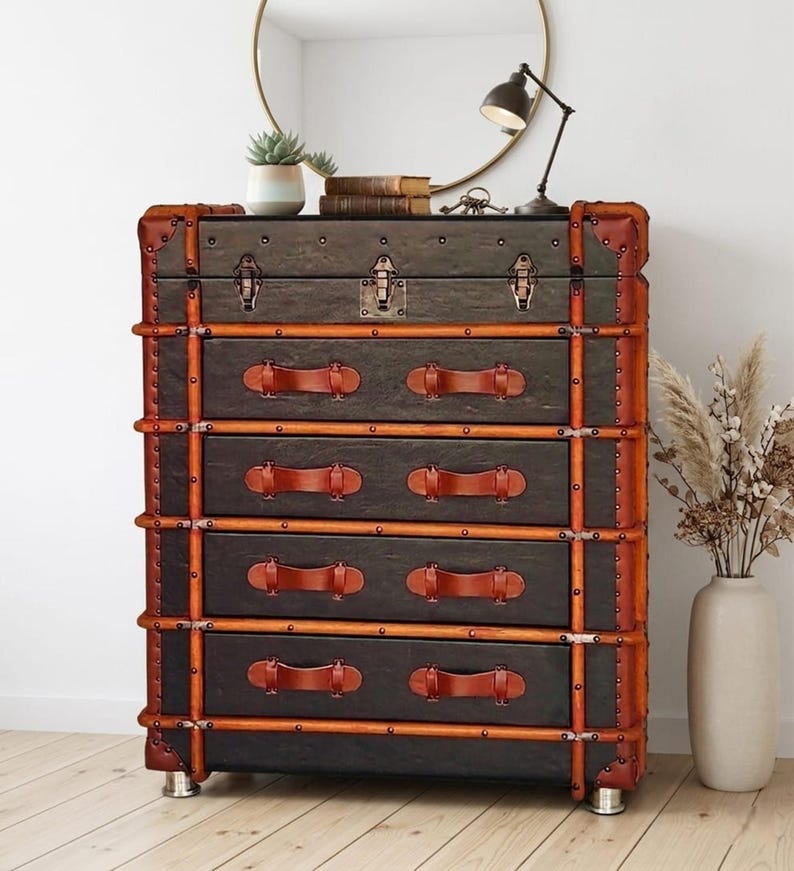 May include: A dark gray chest of drawers with five drawers, trimmed with brown leather and metal accents. The chest has brown leather handles and metal corner protectors. The chest is on silver metal feet.