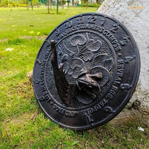 Könnte beinhalten: Eine schwarze Sonnenuhr aus Gusseisen mit einem Frosch- und Seerosen-Design. Die Sonnenuhr hat römische Ziffern und den Text "A Quick Summers Glimpse Reveal Egg, Tadpole, and Frog. Life in Constant Change."