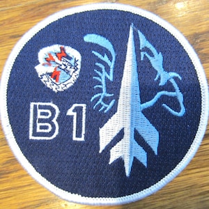 May include: A blue and white embroidered patch with a white B1 and a stylized blue and white rocket design. The rocket is surrounded by a blue and white stylized design.