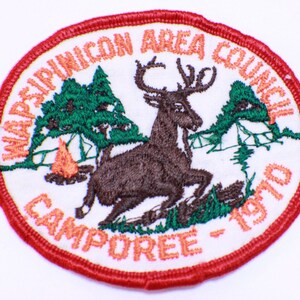May include: A white embroidered patch with a red border. The patch features a brown deer running through a green forest with a campfire in the background. The text "Wapsinoncon Area Council Camporee - 1970" is embroidered in orange around the edge of the patch.