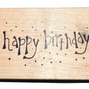 May include: Wooden stamp with the text "happy birthday" in a whimsical font with black ink. The stamp is surrounded by small black dots.
