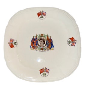 May include: A white ceramic plate with a red, white, and blue design commemorating the coronation of Queen Elizabeth II. The plate features a portrait of the Queen, the British royal coat of arms, and crossed flags of the United Kingdom and Canada.