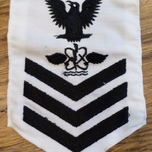 May include: White fabric patch with black embroidered insignia. The insignia features a stylized eagle with wings spread, a medical caduceus, and three black chevrons.