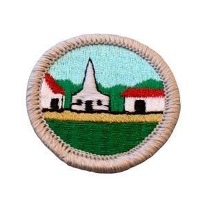 May include: Embroidered patch featuring a quaint village scene. The design includes a white church with a steeple, two houses with red roofs, and green grass under a light blue sky. The patch is round with a tan border.