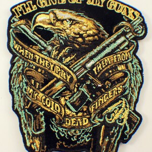 May include: A black embroidered patch with a gold eagle holding two guns crossed. The eagle is surrounded by a gold ribbon with the text "I'll give up my guns when they pry them from my cold dead fingers".