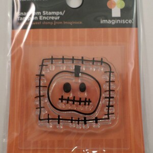 May include: Clear plastic stamp with a black outline of a stitched pumpkin with a face. The stamp is packaged on a card with the text "Spooky Town Collection Pumpkin Patch Stamp/1 Tampon" and "Imaginisce" in black text.