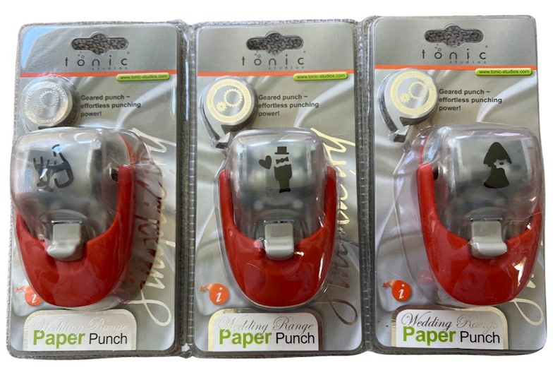 K&ouml;nnte beinhalten: Drei rote und silberne Papierstanzer mit durchsichtigen Plastikdeckeln. Jeder Stanzer zeigt ein anderes Design zum Thema Hochzeit: ein Brautpaar, ein Brautpaar mit einem Herzen und eine Braut mit Schleier. Die Verpackung ist silberfarben mit dem Wort "Tonic" in Schwarz gedruckt.