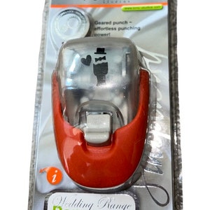 K&ouml;nnte beinhalten: Ein roter und silberner Papierstanzer mit Hochzeitsthema. Der Stanzer zeigt einen Br&auml;utigam mit Zylinder und einem Herzen. Die Verpackung lautet "Wedding Range Paper Punch".