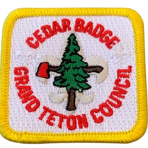 May include: A white embroidered patch with a yellow border. The patch features a green pine tree with a brown trunk and a red axe. The text "CEDAR BADGE GRAND TETON COUNCIL" is in red around the tree.