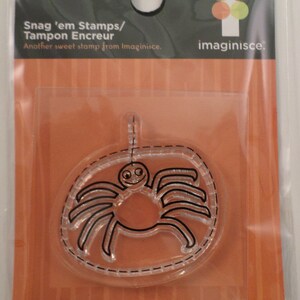 May include: A clear spider-shaped stamp from the Spooky Town Collection, packaged in a clear plastic sleeve. The stamp features a smiling spider design with black lines and the text "Snag 'em Stamps/Tampon Encreur".