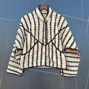 May include: A long-sleeve quilted jacket in cream with a repeating black triangular pattern. The jacket features a high collar, a front button closure with four visible buttons, and black trim along the edges, seams, and two front pockets.
