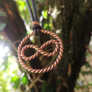 May include: A copper-colored infinity symbol pendant, crafted from twisted wire, hangs from a dark cord. The pendant is suspended against a blurred background of green foliage and tree bark, creating a natural aesthetic.