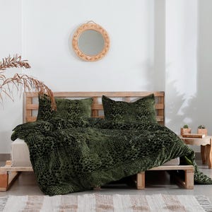 K&ouml;nnte beinhalten: Ein Bett mit einer gr&uuml;n gemusterten Bettdecke und passenden Kissen. Das Bett ist aus Holz und hat eine Matratze in neutraler Farbe. Ein dekorativer Spiegel h&auml;ngt an der Wand &uuml;ber dem Bett. Eine hohe Vase mit getrockneten Pflanzen und eine Topfpflanze befinden sich ebenfalls im Raum.