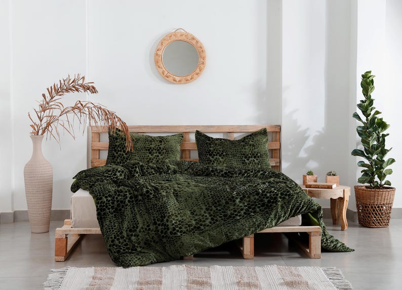 K&ouml;nnte beinhalten: Ein Bett mit einer gr&uuml;n gemusterten Bettdecke und passenden Kissen. Das Bett ist aus Holz und hat eine Matratze in neutraler Farbe. Ein dekorativer Spiegel h&auml;ngt an der Wand &uuml;ber dem Bett. Eine hohe Vase mit getrockneten Pflanzen und eine Topfpflanze befinden sich ebenfalls im Raum.