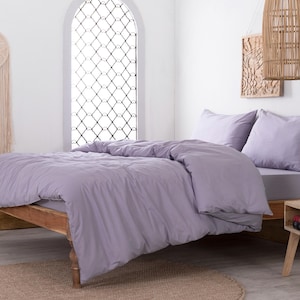 May include: A bedroom with a bed dressed in lavender bedding. The wooden bed frame is on a round rug. A decorative dream catcher hangs on the wall. A side table holds a vase and books, with a woven lampshade overhead.