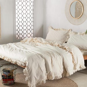 May include: Cream-colored bedding set with ruffled edges on a wooden bed frame. The room features a decorative wall hanging, a round mirror, and a small side table. A metal basket with books sits on a woven rug.