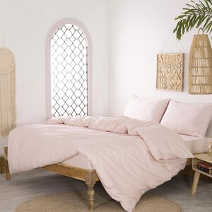May include: A bedroom scene featuring a bed with light pink bedding, including a duvet and pillows. The room has a wooden bed frame, a small wooden side table, and decorative wall hangings. A woven basket with dried plants is on the floor.