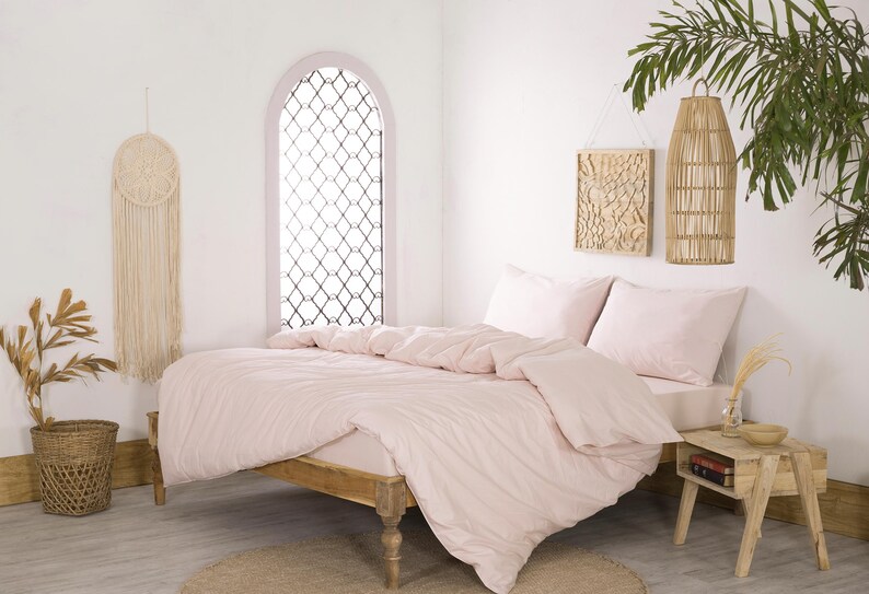 May include: A bedroom scene featuring a bed with light pink bedding, including a duvet and pillows. The room has a wooden bed frame, a small wooden side table, and decorative wall hangings. A woven basket with dried plants is on the floor.