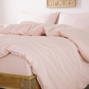 May include: A bed with a light pink duvet cover and matching pillowcases. The bed frame is made of light-colored wood. The bedding is neatly arranged, creating a soft and inviting look. The room has a minimalist aesthetic.