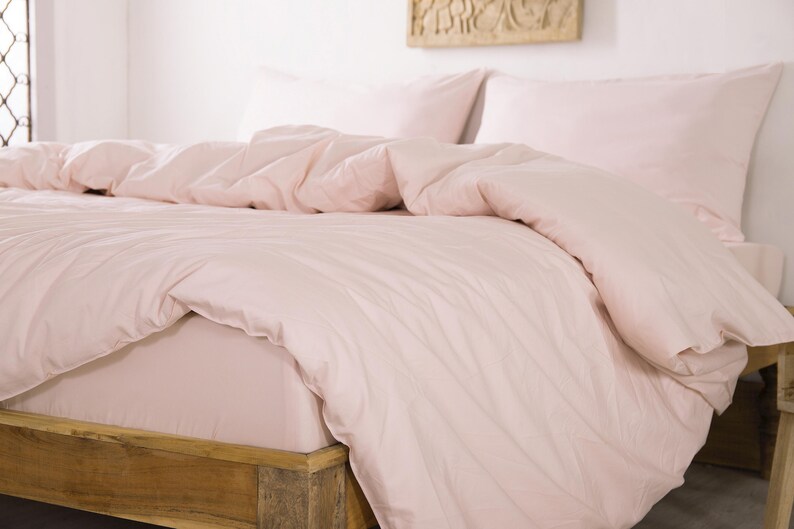 May include: A bed with a light pink duvet cover and matching pillowcases. The bed frame is made of light-colored wood. The bedding is neatly arranged, creating a soft and inviting look. The room has a minimalist aesthetic.