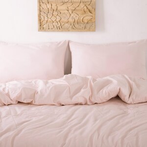 May include: A bed with light pink bedding, including two pillowcases and a comforter. The bedding is neatly arranged, creating a soft and inviting look. A decorative wooden panel hangs on the wall above the bed.