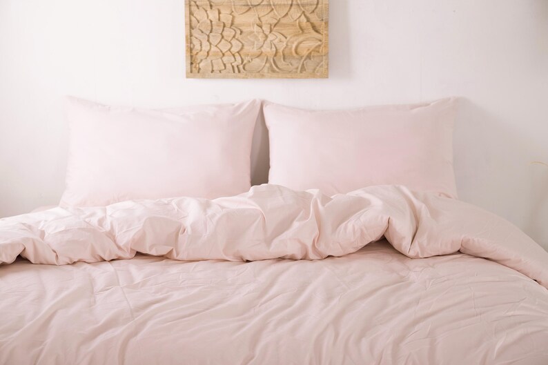 May include: A bed with light pink bedding, including two pillowcases and a comforter. The bedding is neatly arranged, creating a soft and inviting look. A decorative wooden panel hangs on the wall above the bed.